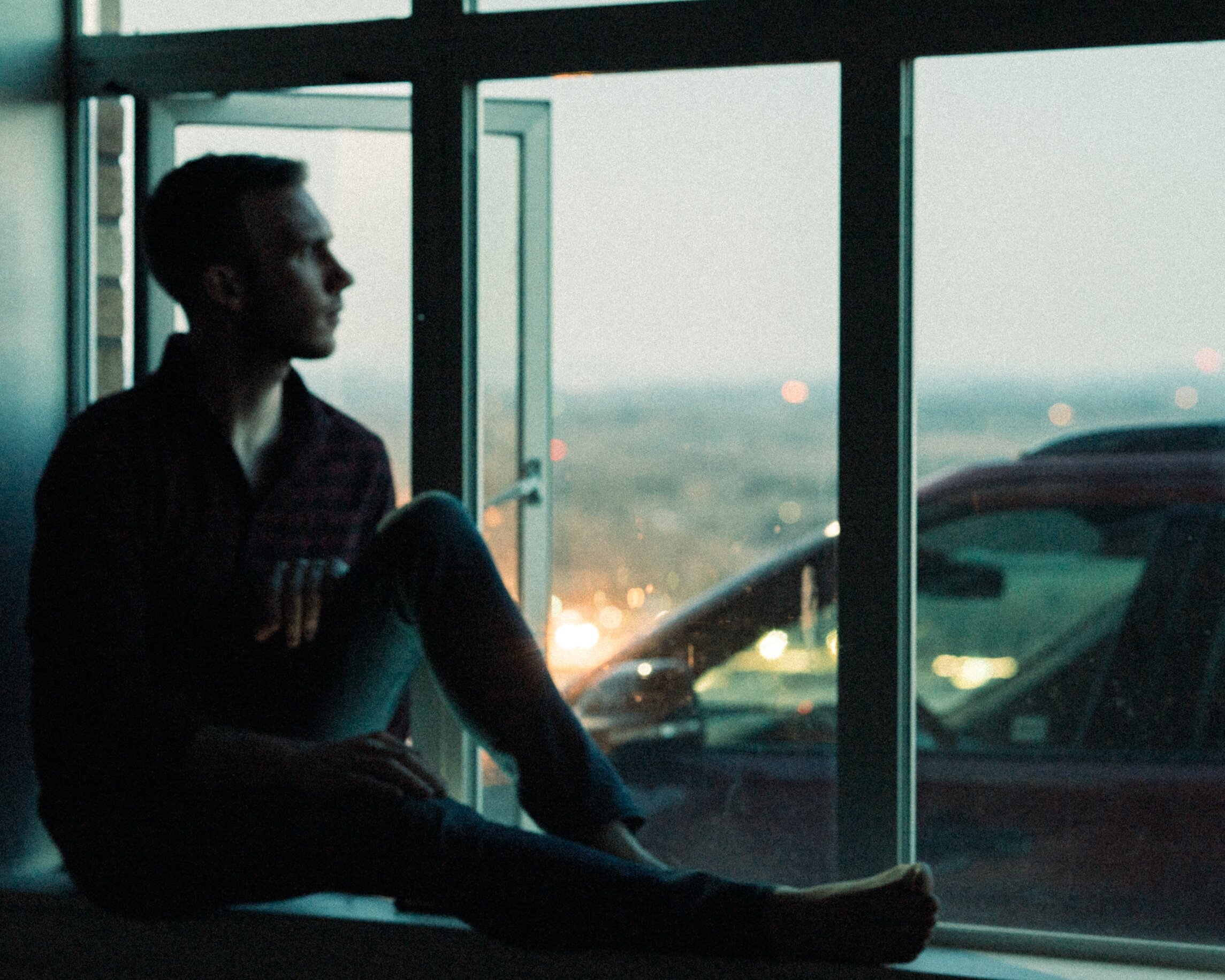 Man sitting on a window sill at dusk staring out of the window
