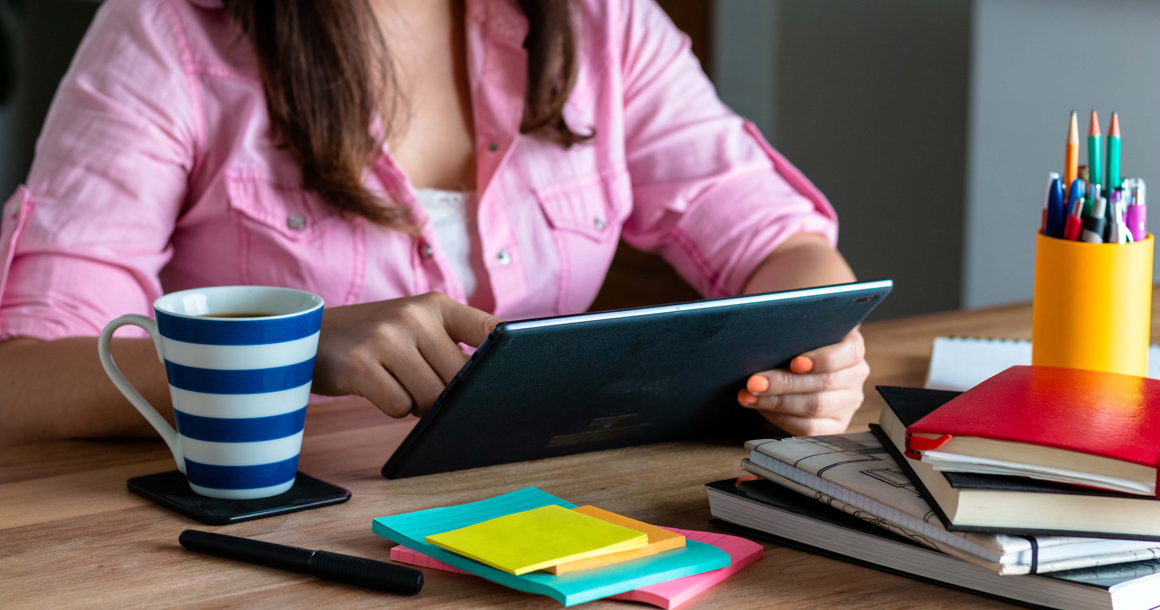 Person sitting at a table using their tablet to do research with a cup of tea next to them. There are pens, stack of post-its and books on the table