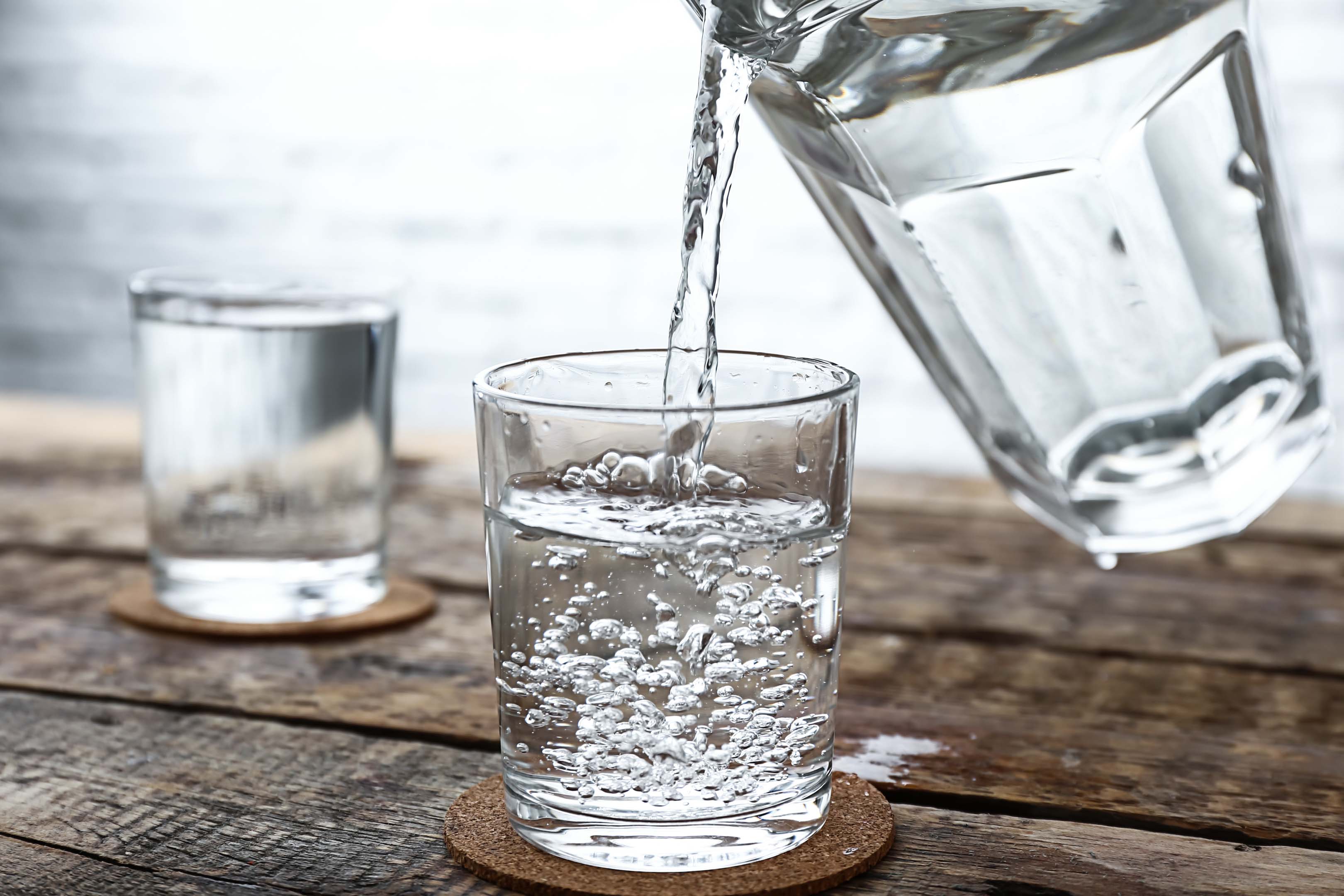 A small glass being filled with water from a clear water jug. The glass is sitting on a brown coaster on a wooden table, with another in the background.