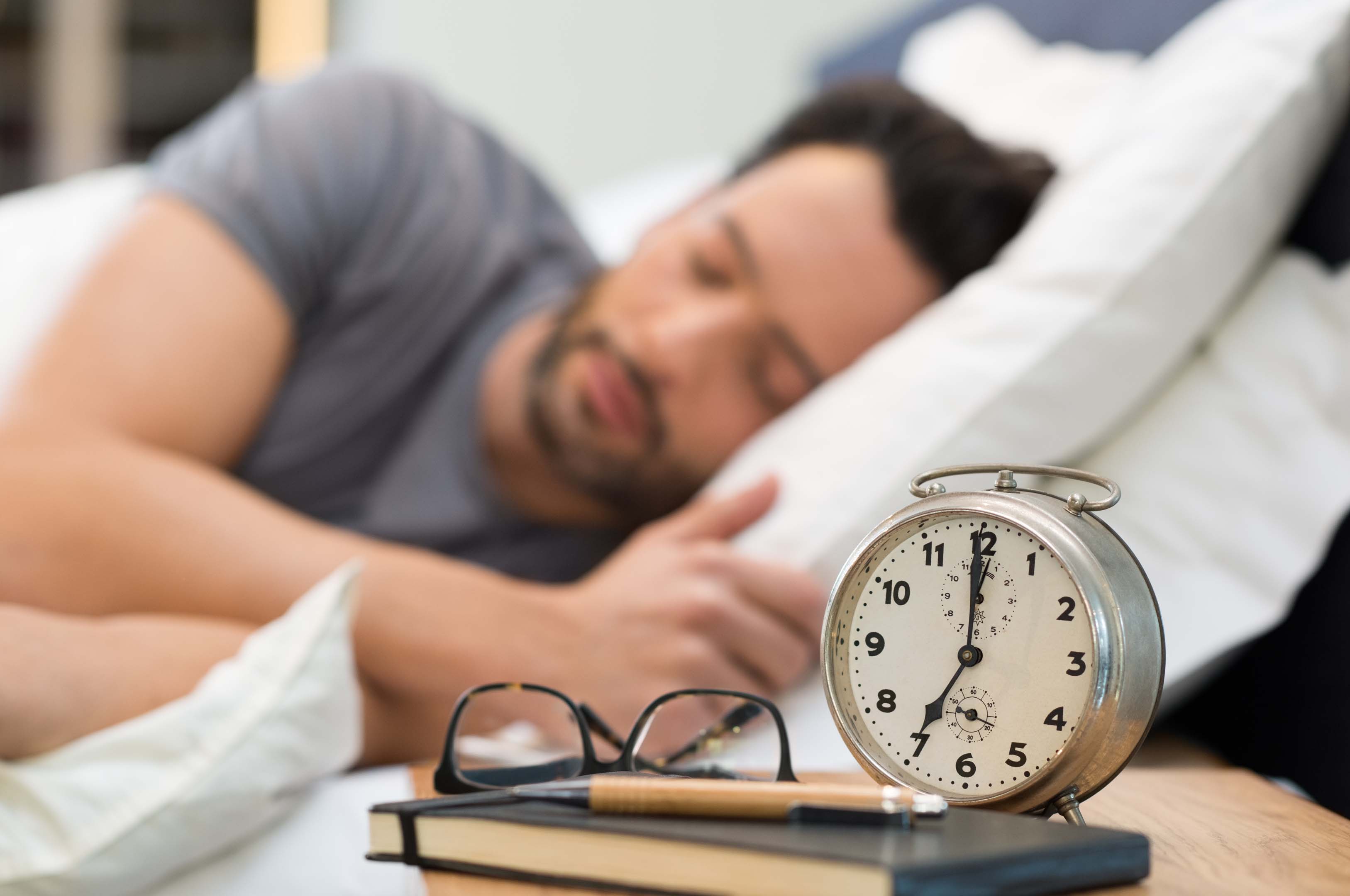 Person asleep with an alarm clock, spectacles and a notepad & pen on a bedside table. Writing down thoughts before bed can help you sleep better