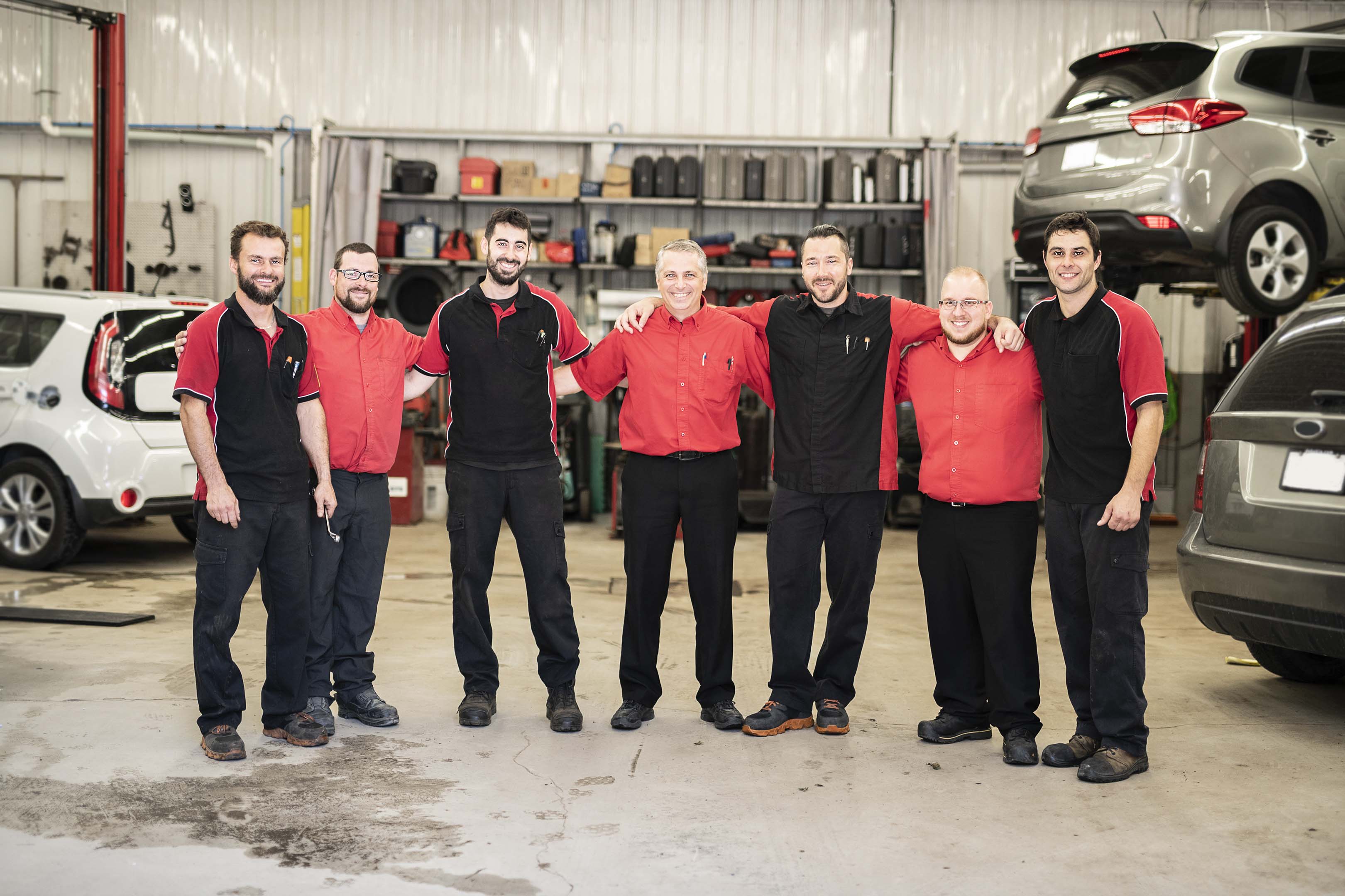 Seven people working in a garage standing in a line with their arms around each other smiling