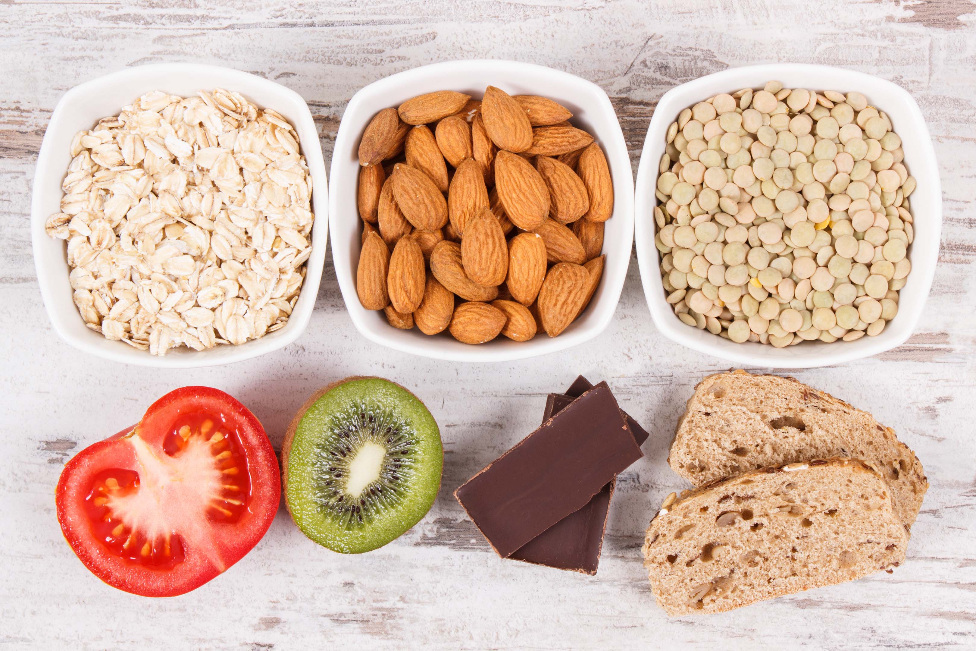 Lots of healthy food is laid out on a wooden table, including bowls of different nuts, fruit, vegetables, dark chocolate and brown bread.