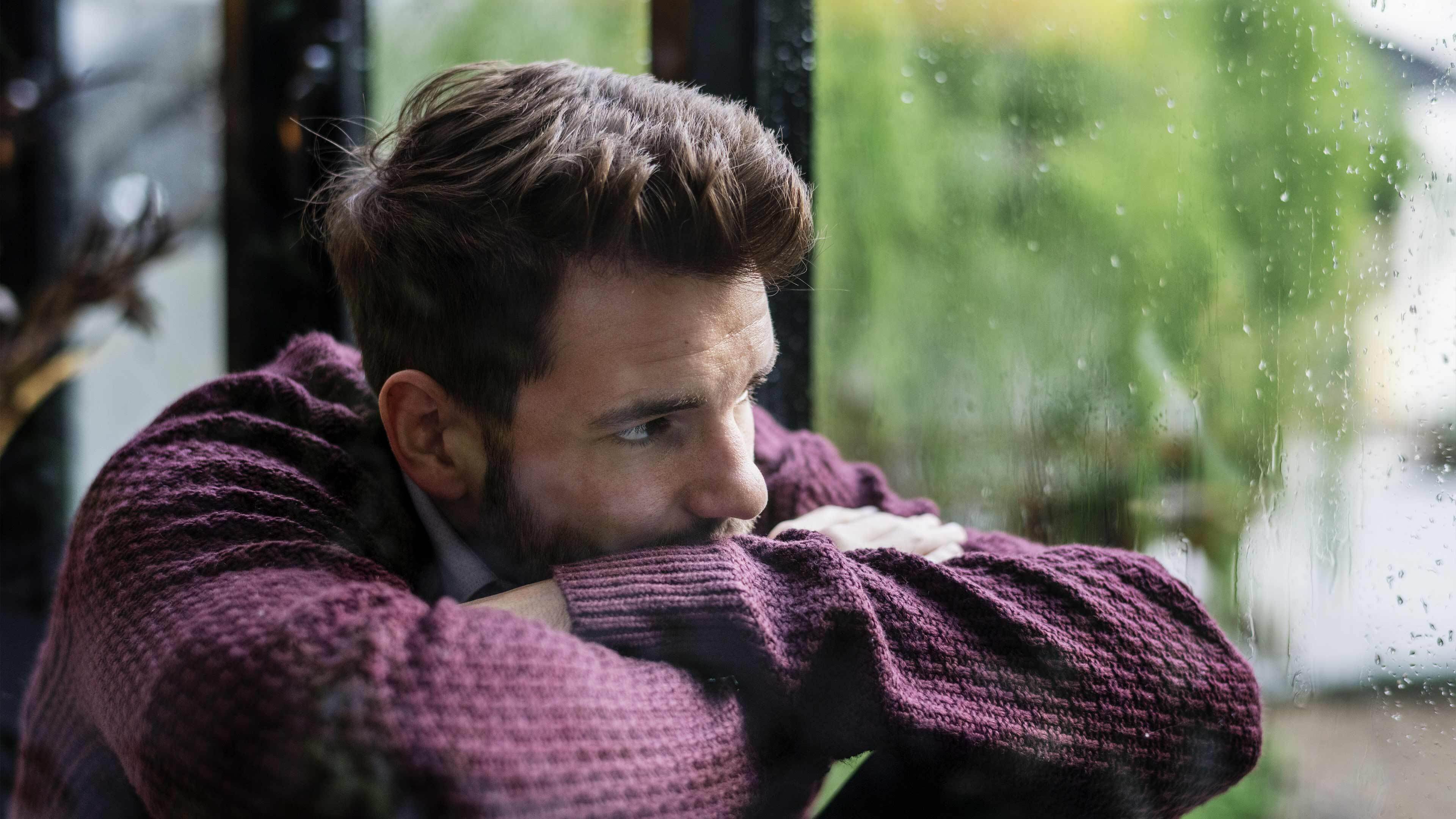 Person resting their chin on crossed arms looking out a window on a rainy day