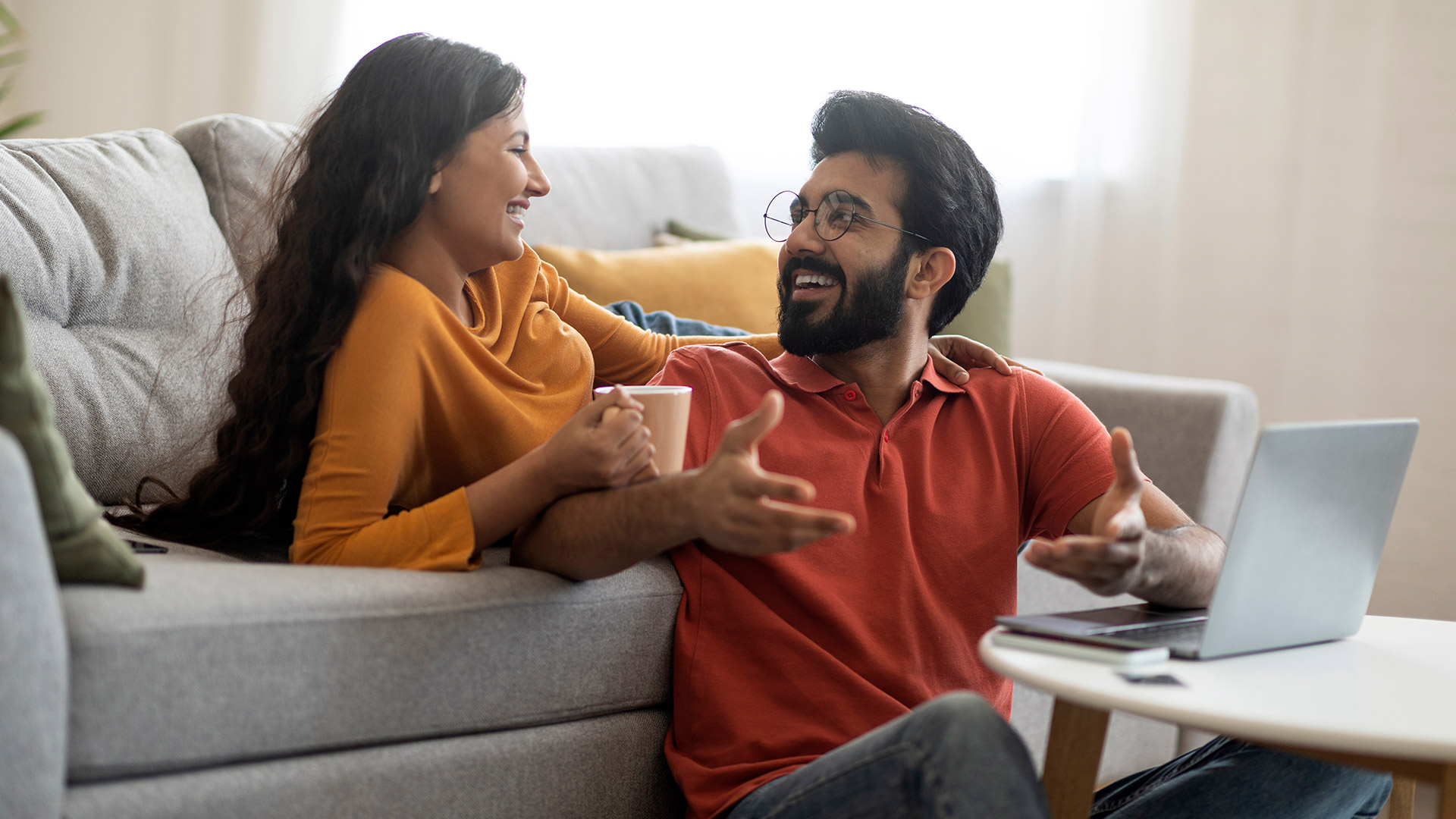 Happy couple discussing finances together on grey sofa with a laptop and coffee at home.