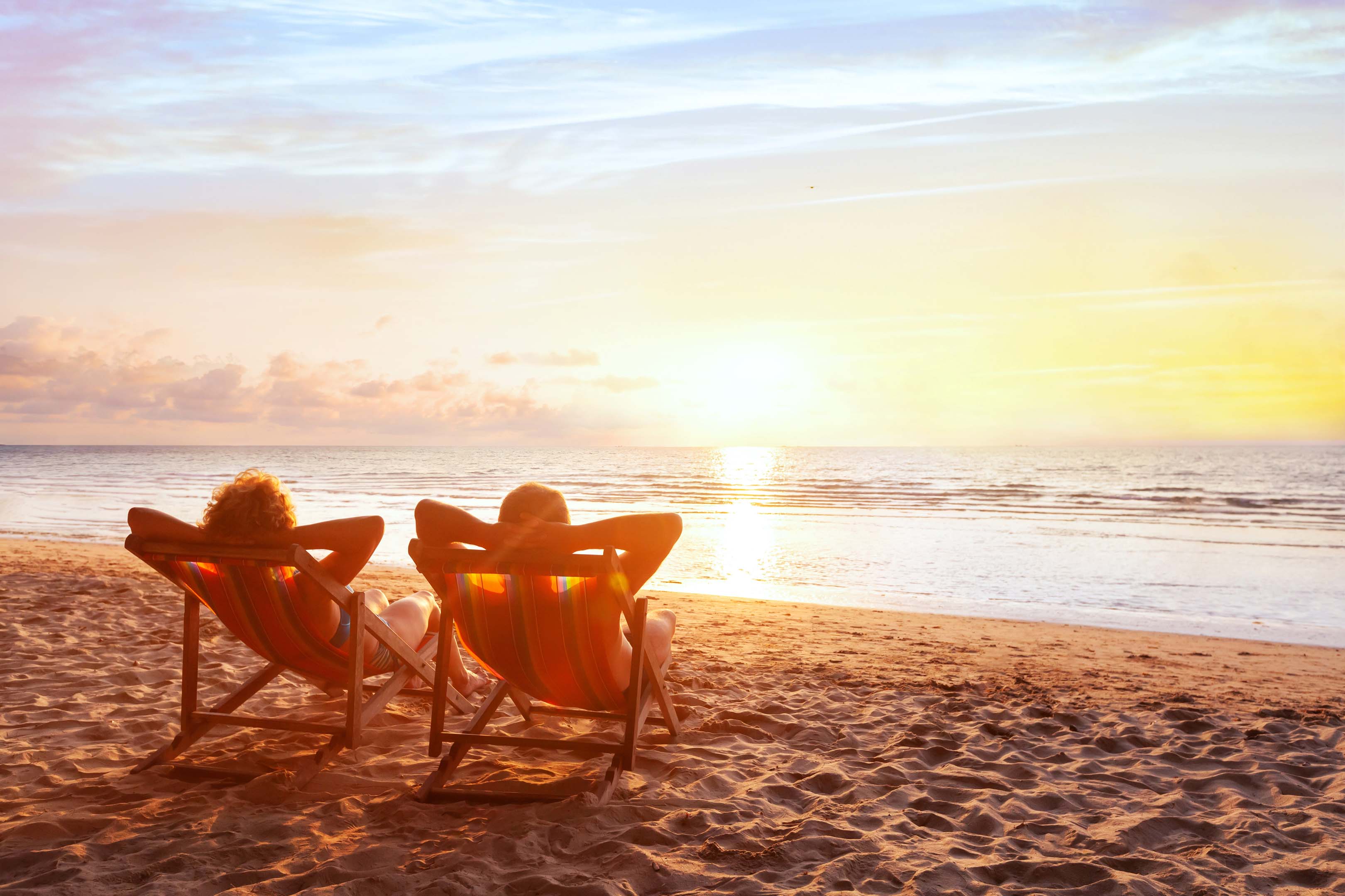 Two people enjoying their retirement sitting on deckchairs on a beach, watching the sunset