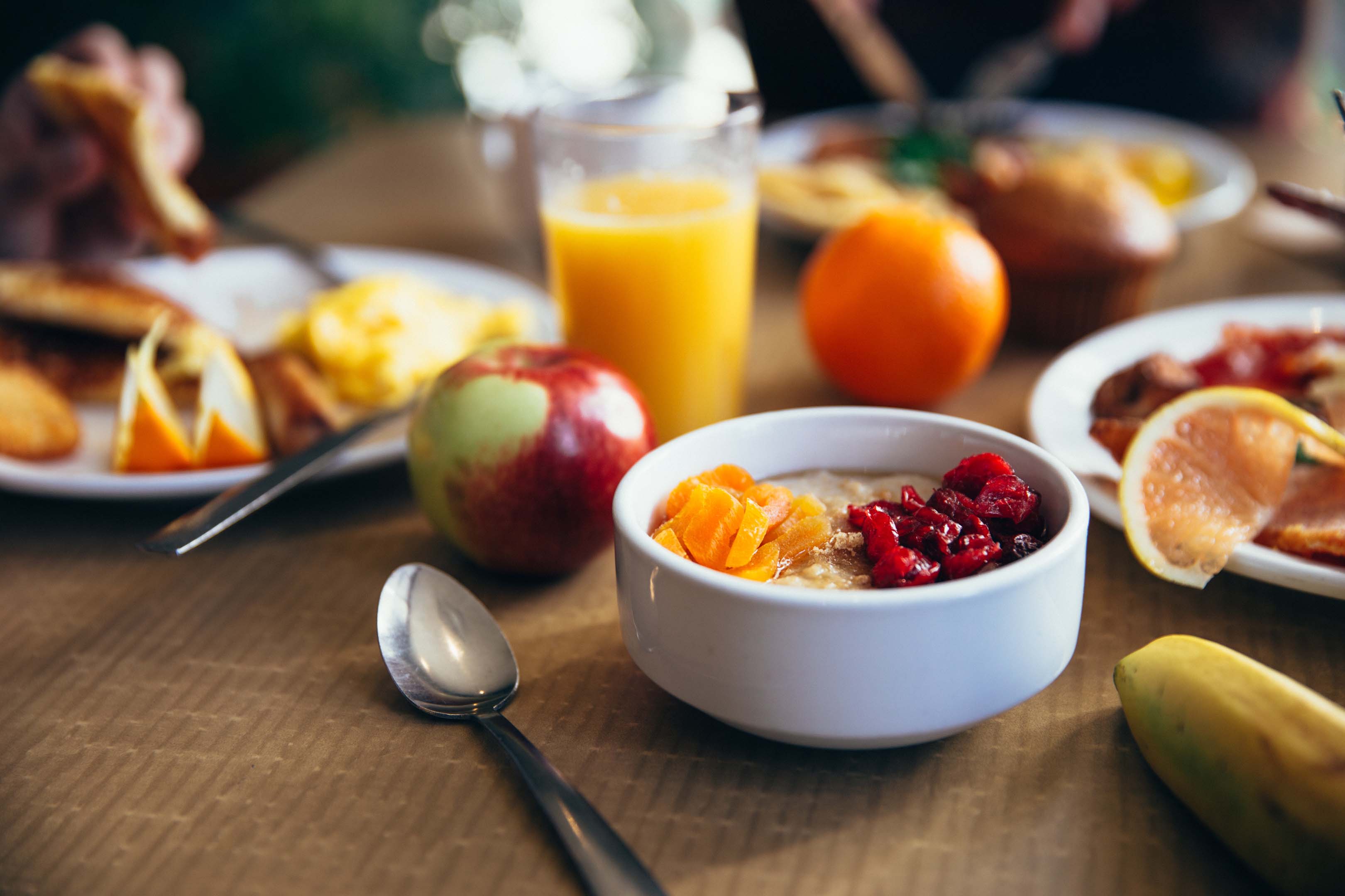 A white bowl full of fruit with a spoon next to it sitting on a table. The rest of the table contains more fruit and other healthy breakfast foods  